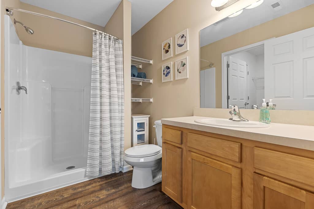 Bathroom with a walk-in shower, striped curtain, toilet, wooden vanity with single sink, wall shelves with towels, and decorative art above the toilet.