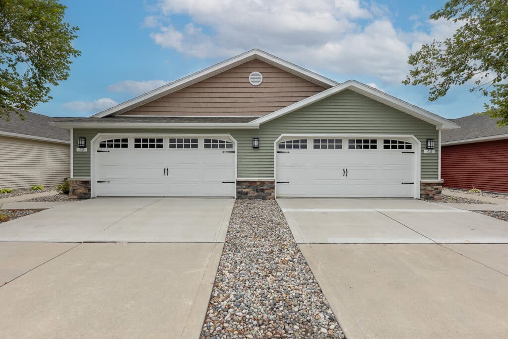A duplex home with two white double garages, green and brown siding, stone accents, and a central rock landscaping strip between driveways.