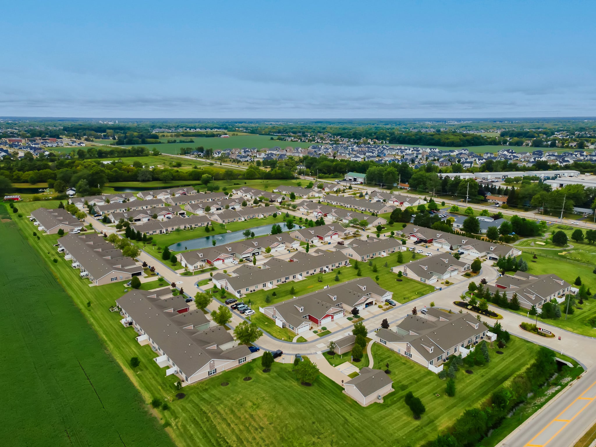 Aerial view of a suburban residential neighborhood with rows of houses, green lawns, roads, and surrounding fields under a partly cloudy sky.