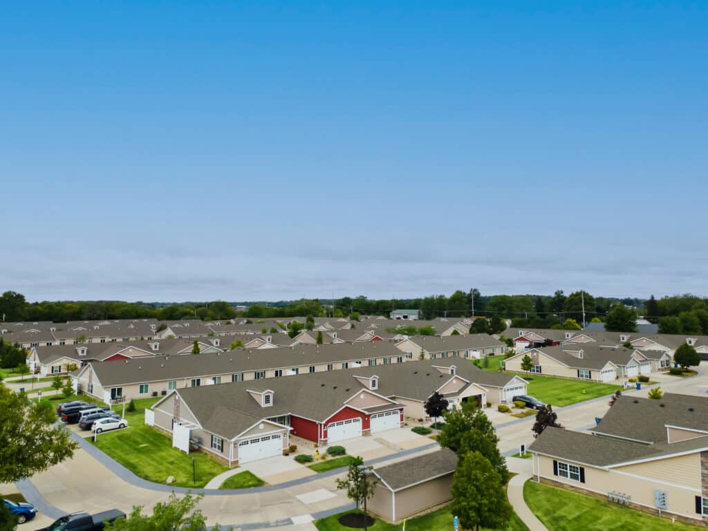 Aerial view of a suburban neighborhood with rows of single-story houses, neatly mowed lawns, driveways, and trees under a clear blue sky.