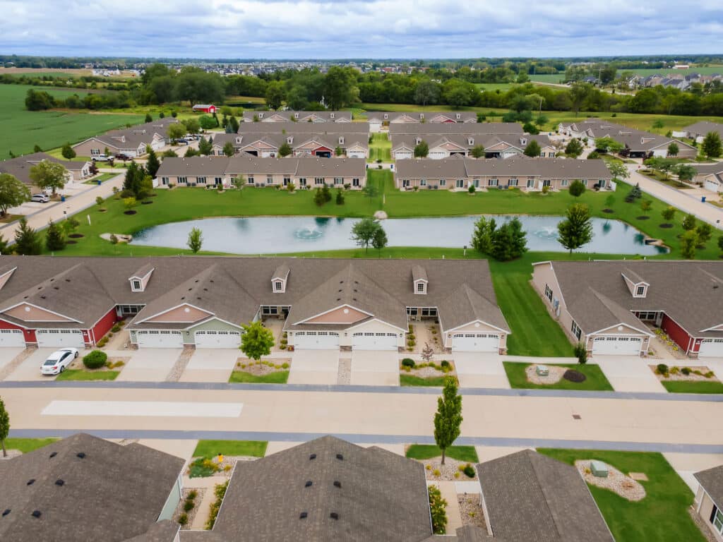Aerial view of a suburban residential neighborhood with rows of houses, green lawns, and a small pond at the center, surrounded by trees and open fields.