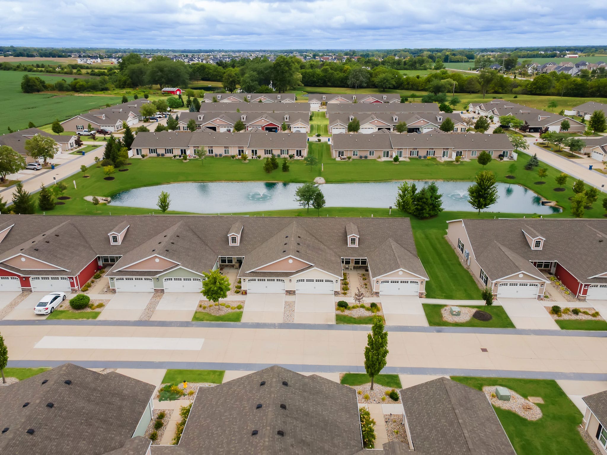 Aerial view of suburban townhomes with attached garages surrounding a small central pond, green lawns, and trees, with fields and distant houses in the background.