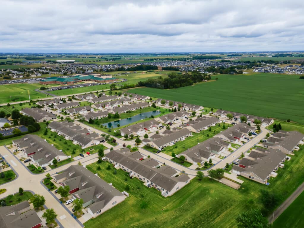 Aerial view of a suburban neighborhood with rows of houses, green lawns, a pond, and surrounding farmland under a cloudy sky.