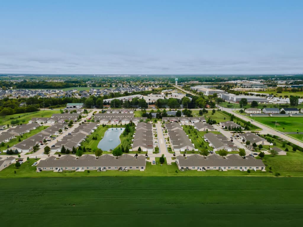 Aerial view of a suburban residential complex with rows of houses, a small pond, roads, and green lawns, surrounded by more neighborhoods and open land.