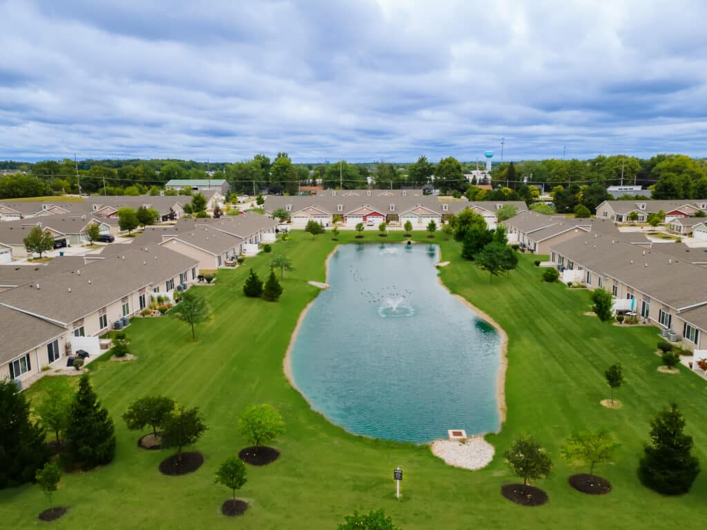 Aerial view of a residential neighborhood with rows of houses surrounding a central pond, green lawns, and scattered trees under a cloudy sky.