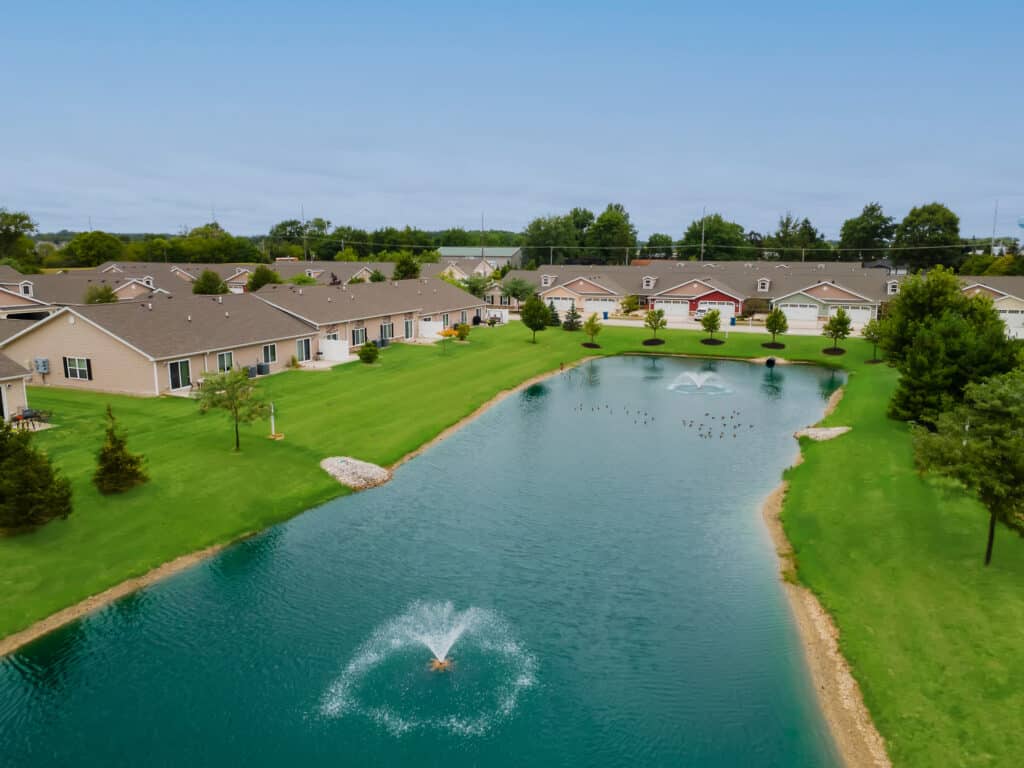 A row of single-story homes lines a pond with a central fountain, surrounded by well-kept green lawns and scattered trees under a clear sky.