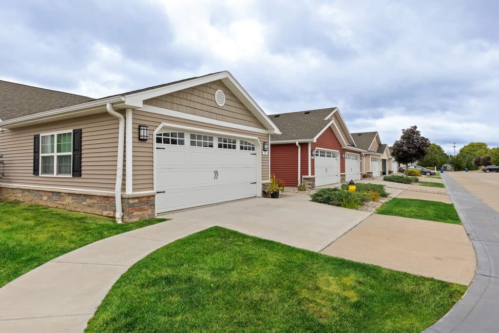Row of single-story suburban houses with attached garages, beige and red siding, neatly trimmed lawns, and a paved sidewalk along the street under a cloudy sky.