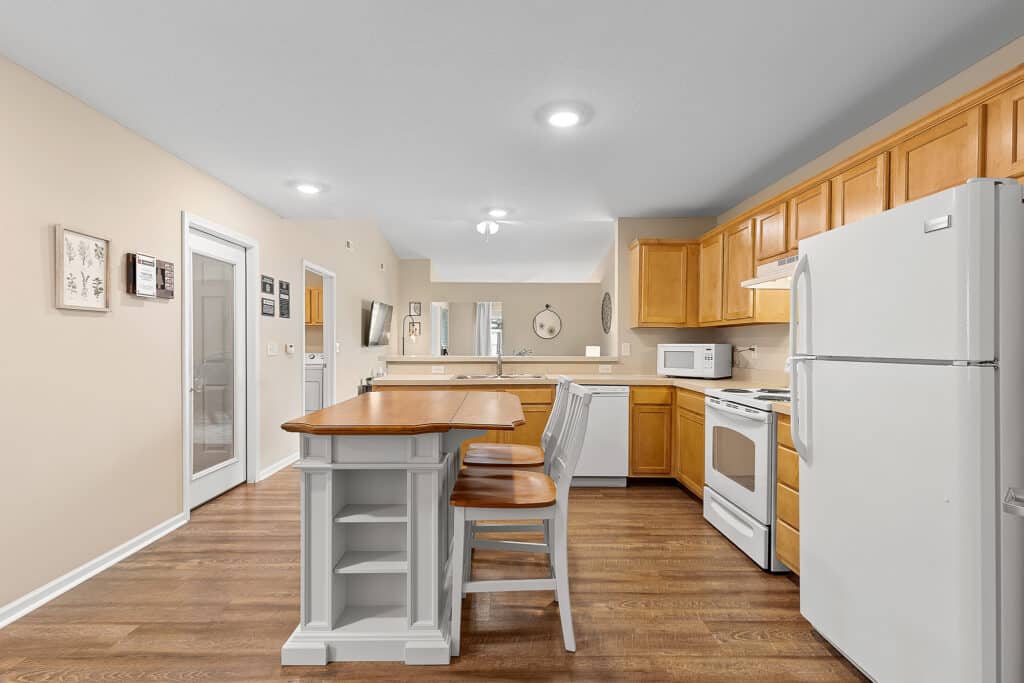 Modern kitchen with wood cabinets, white appliances, an island with seating, and hardwood floors. A dining area and living space are visible in the background.
