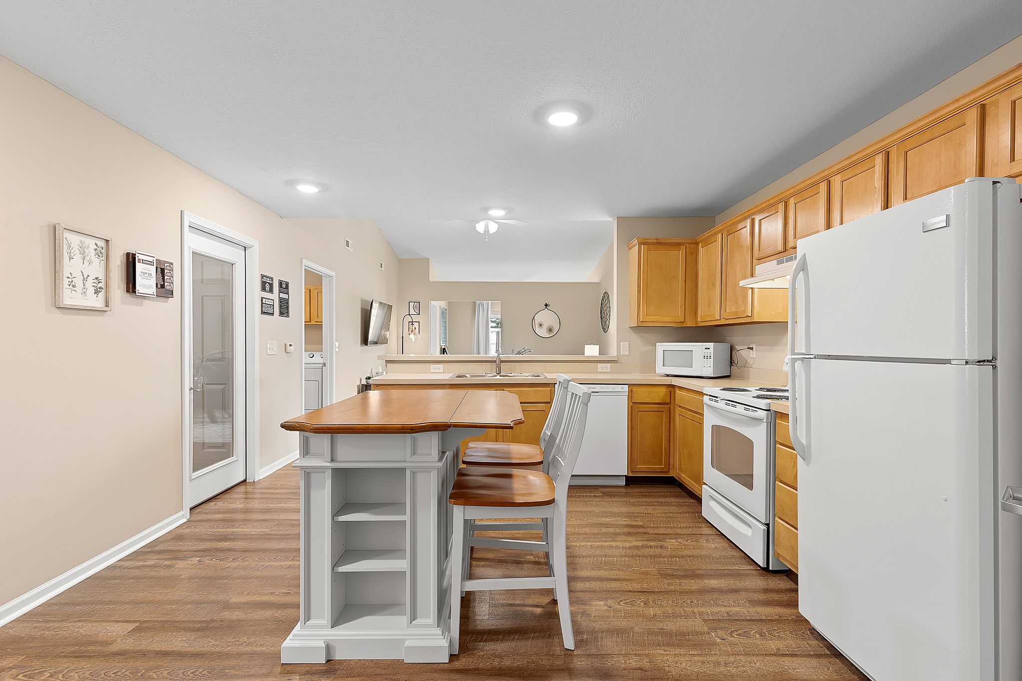 Modern kitchen with wood cabinets, white appliances, an island with seating, and hardwood floors. A dining area and living space are visible in the background.