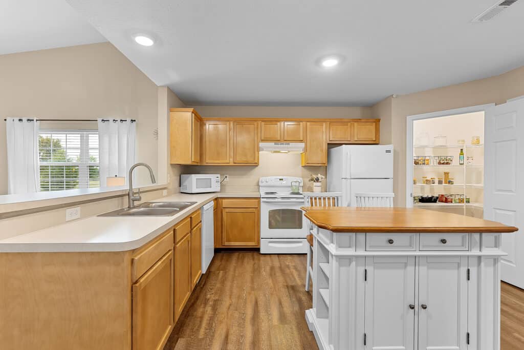 Modern kitchen with wooden cabinets, white appliances, a central island with drawers, visible pantry, and a window with white curtains.
