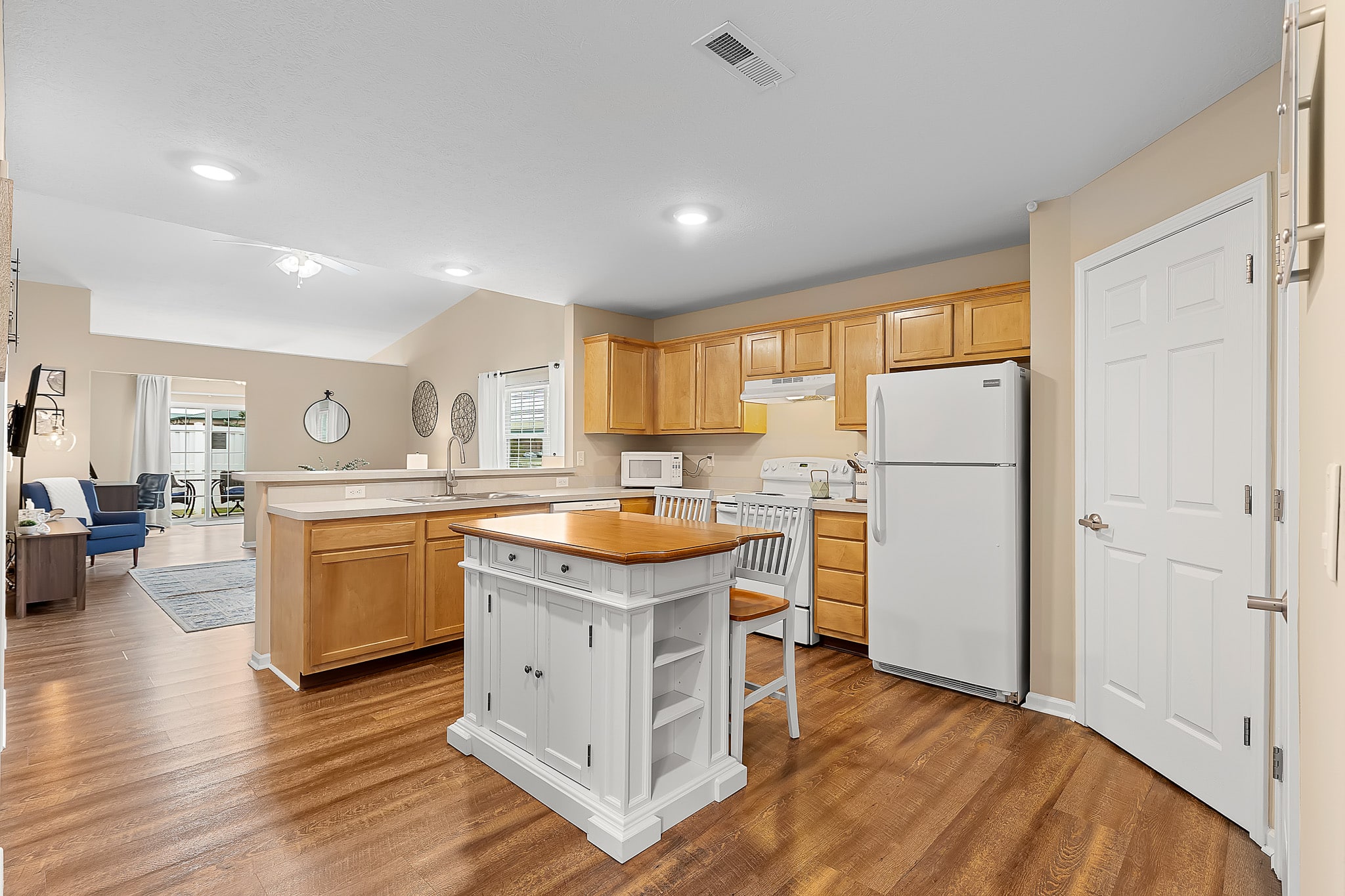 Open kitchen with light wood cabinets, white appliances, a small island with shelving and a chair, and wood-style flooring; living area visible in the background.