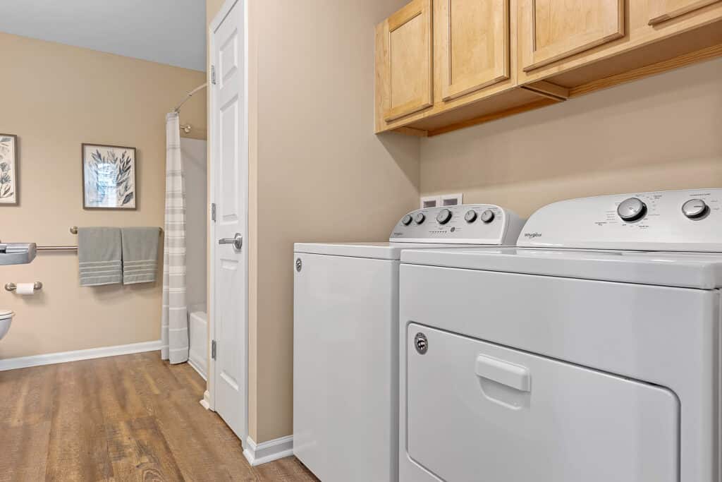 Laundry area with a washing machine and dryer next to a bathroom; wooden cabinets are mounted above, and a bathtub with a shower curtain is visible in the background.