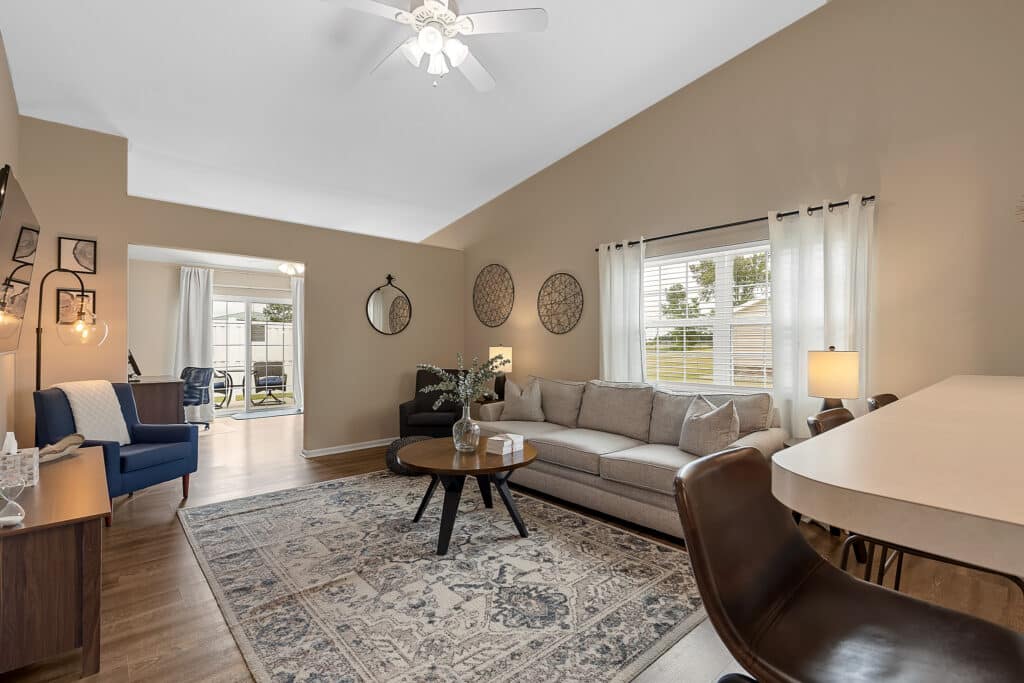 A modern living room with a beige sofa, patterned rug, armchairs, round coffee table, and large windows with white curtains letting in natural light.