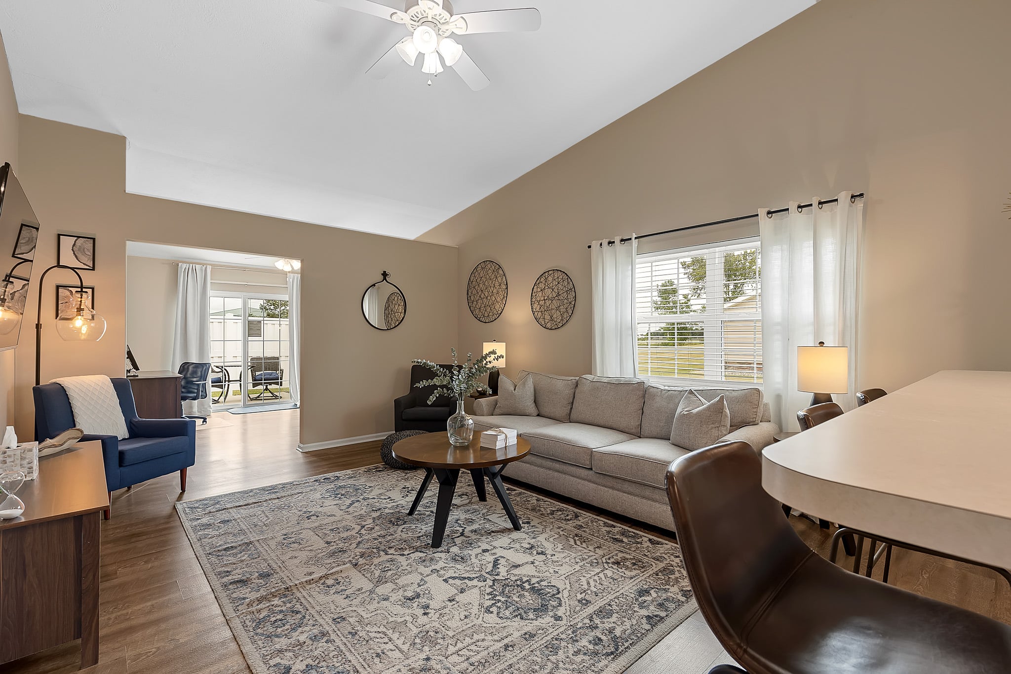 A modern living room with a beige sofa, patterned rug, armchairs, round coffee table, and large windows with white curtains letting in natural light.