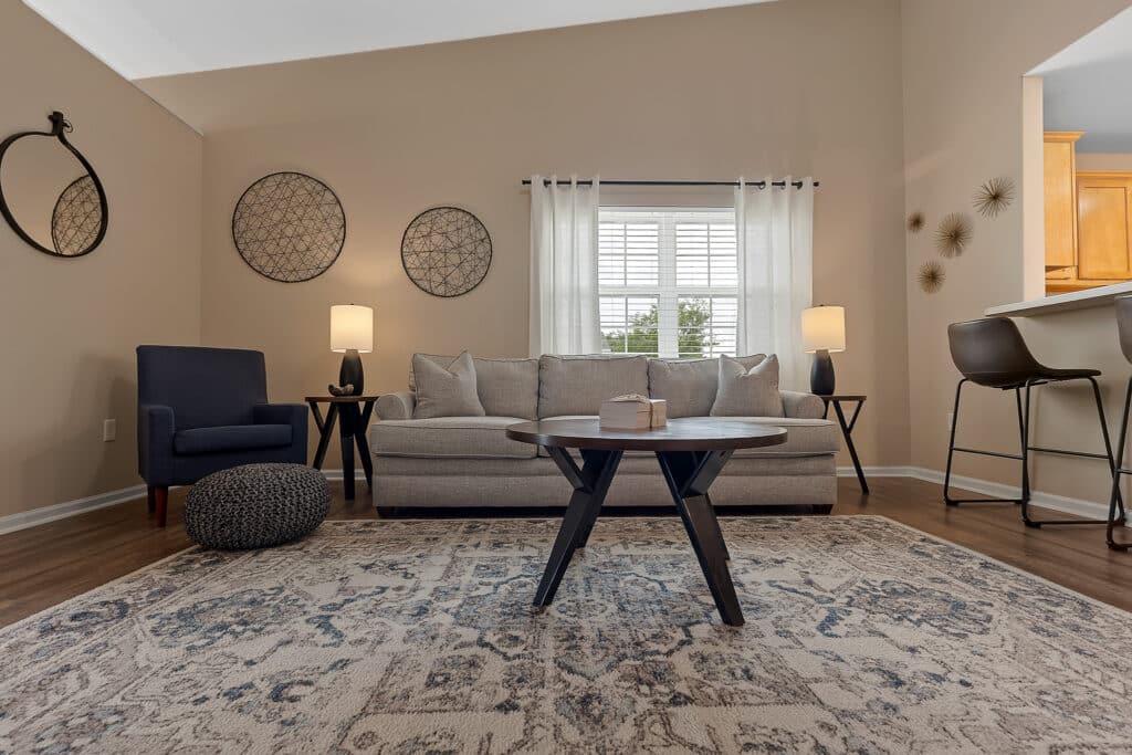 A modern living room with a beige sofa, round coffee table, armchair, and decorative wall art, featuring neutral tones and a patterned rug.