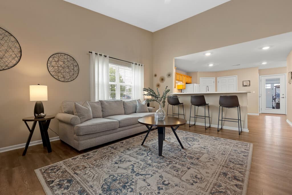 A living room with a beige sofa, round coffee table, patterned rug, bar seating at a counter, and neutral-colored walls. A window with white curtains lets in natural light.