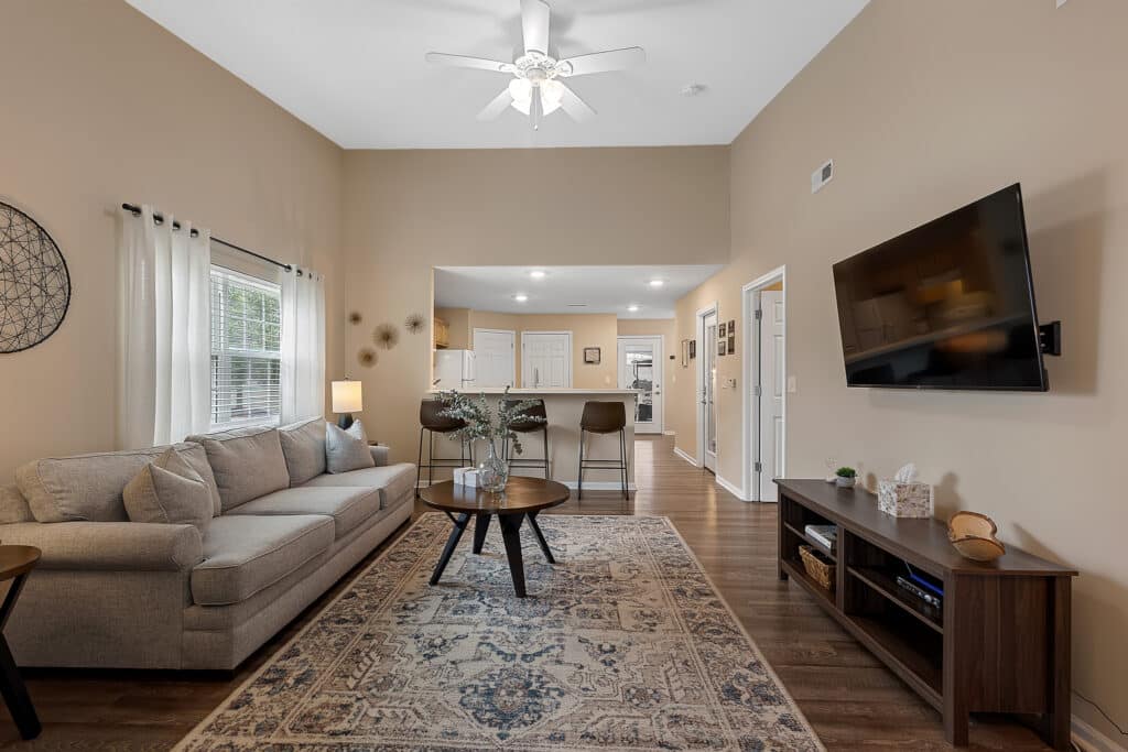 Living room with beige walls, a gray sofa, a patterned rug, a wall-mounted TV, and a view into a kitchen with white cabinets and a breakfast bar.