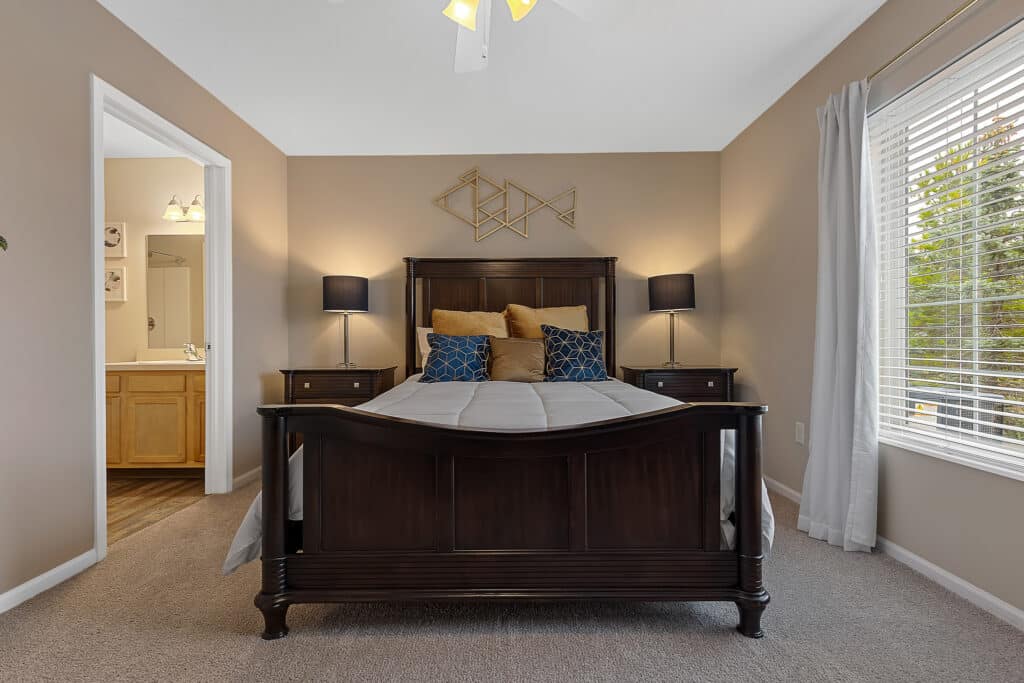 A neatly made bed with dark wood frame, two nightstands with lamps, wall decor above the headboard, and a window with blinds in a beige bedroom; bathroom visible through open door.