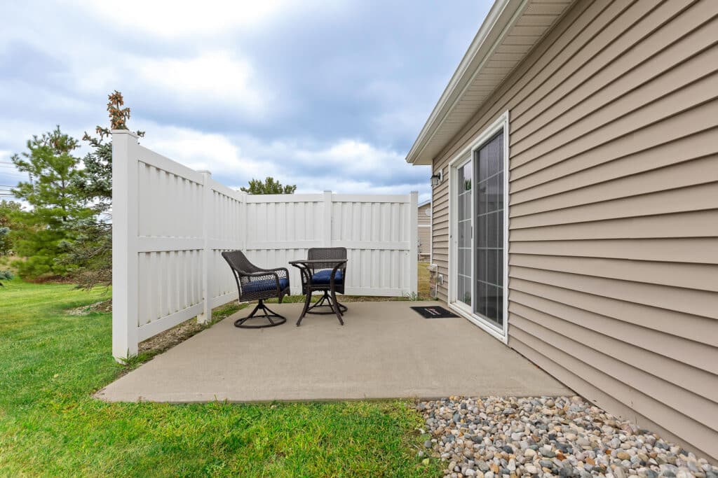 Small concrete patio with two black chairs and a table, next to a beige siding house, white fence, sliding glass door, and landscaped rocks. Grass and trees in the background.