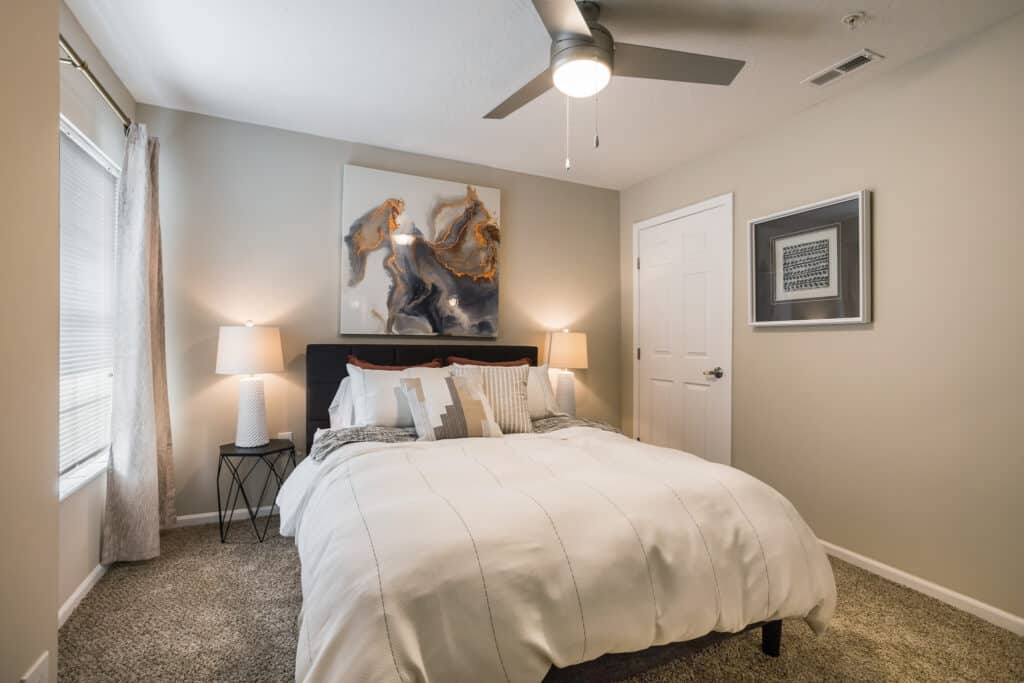 A modern bedroom with a neatly made bed, two bedside tables with lamps, abstract wall art above the headboard, and a ceiling fan.