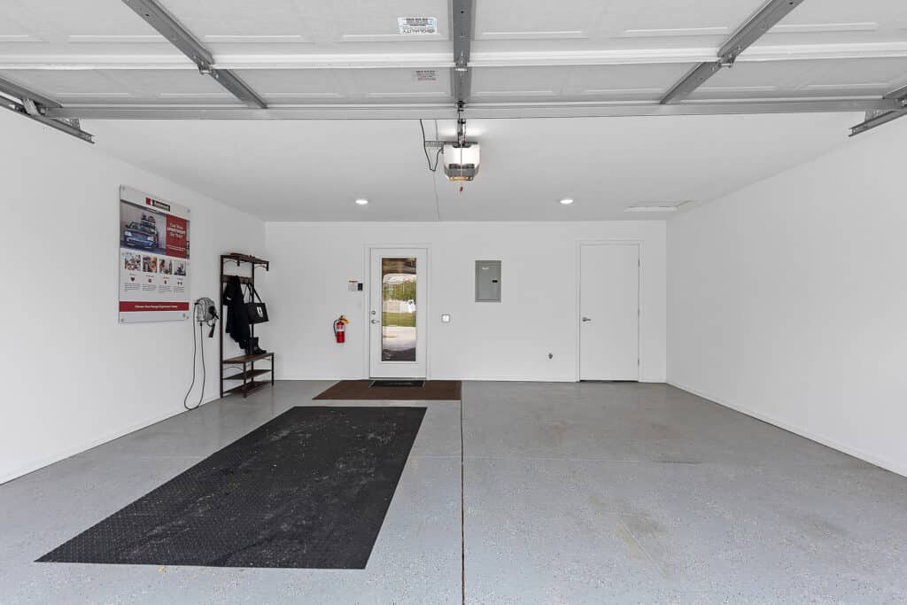 Interior of a clean, empty garage with white walls, a gray speckled floor, a door at the back, wall-mounted shelves, and a garage door opener on the ceiling.