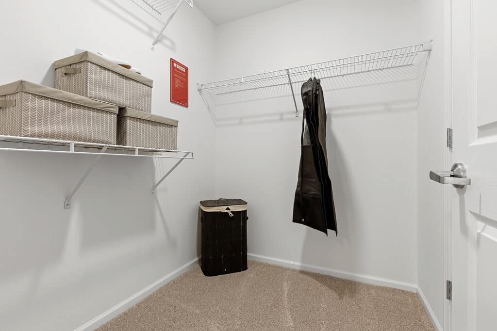 A walk-in closet with carpeted floor, wire shelves holding storage boxes, a hamper, a red sign on the wall, and a pair of pants hanging from the rack.
