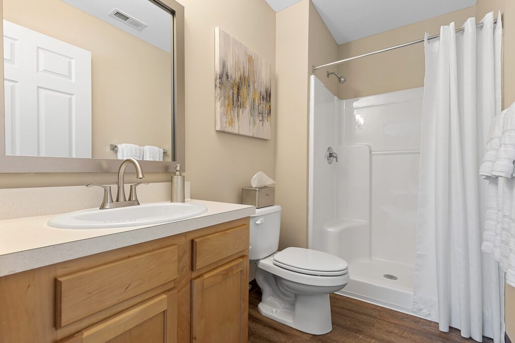 A bathroom with a single sink vanity, a toilet, a walk-in shower with a white curtain, and beige walls decorated with a framed abstract painting.