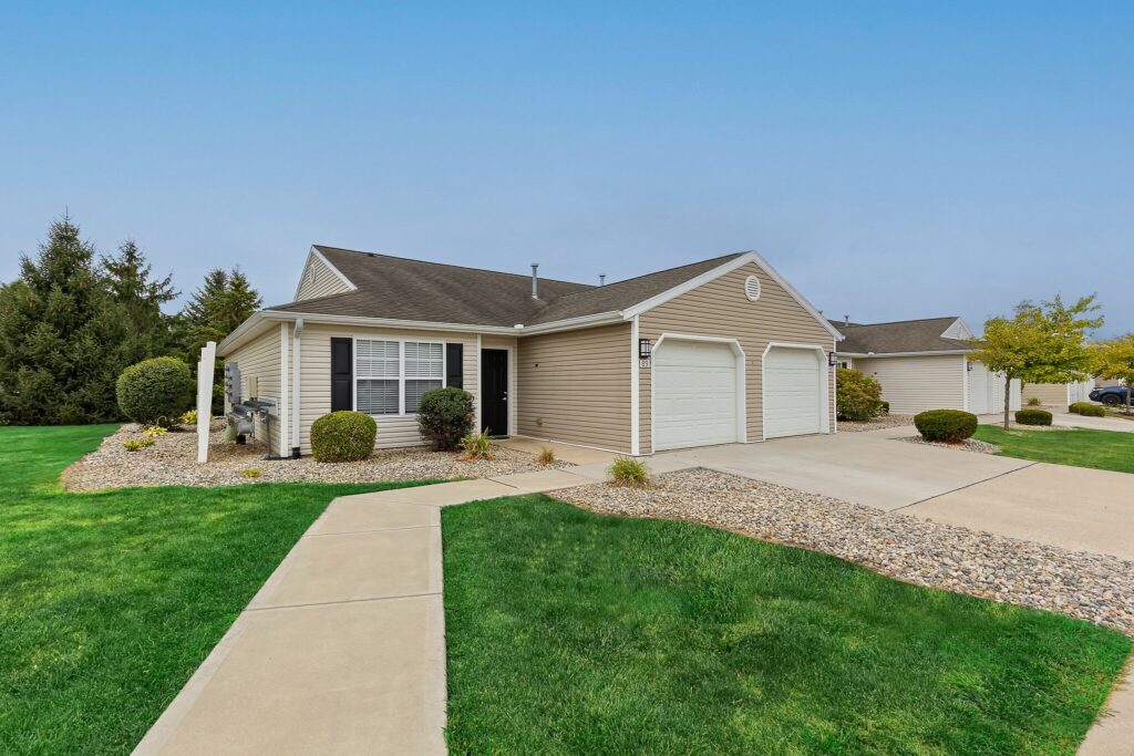 Single-story beige house with white trim, two-car garage, manicured lawn, shrubs, and a concrete walkway leading to the front door on a clear day.