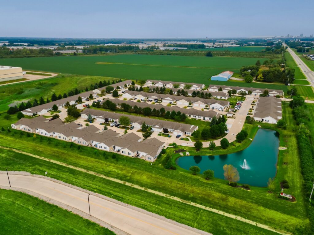 Aerial view of a suburban residential complex surrounded by green fields, featuring rows of single-story homes, a pond with a fountain, and a nearby road.