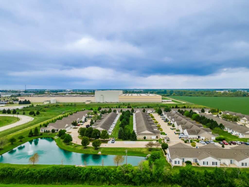 Aerial view of a suburban residential neighborhood with rows of houses, a pond, green fields, and industrial buildings in the background under a cloudy sky.