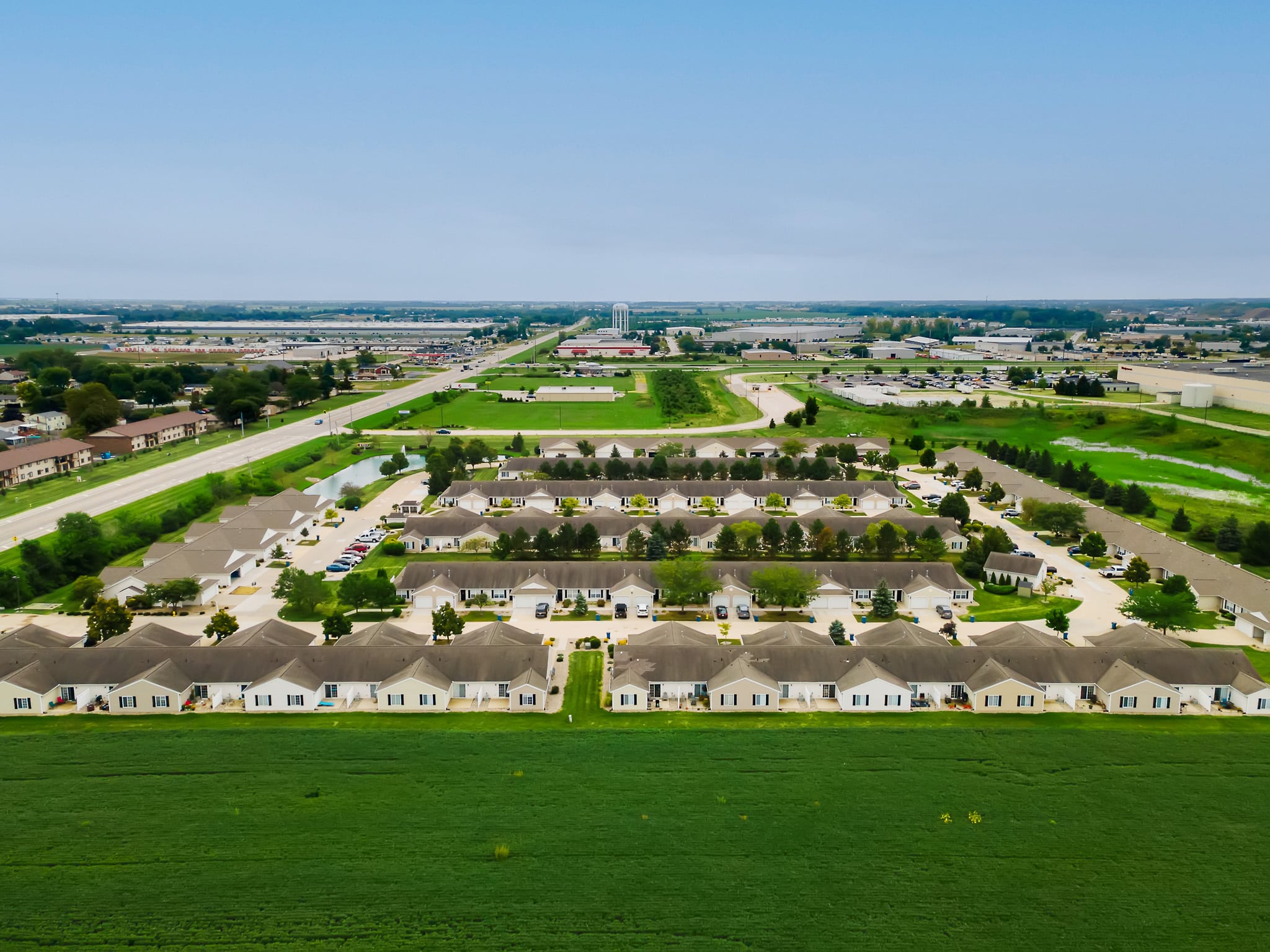 Aerial view of a suburban neighborhood with rows of identical houses, green lawns, and surrounding roads, with commercial buildings visible in the distance.