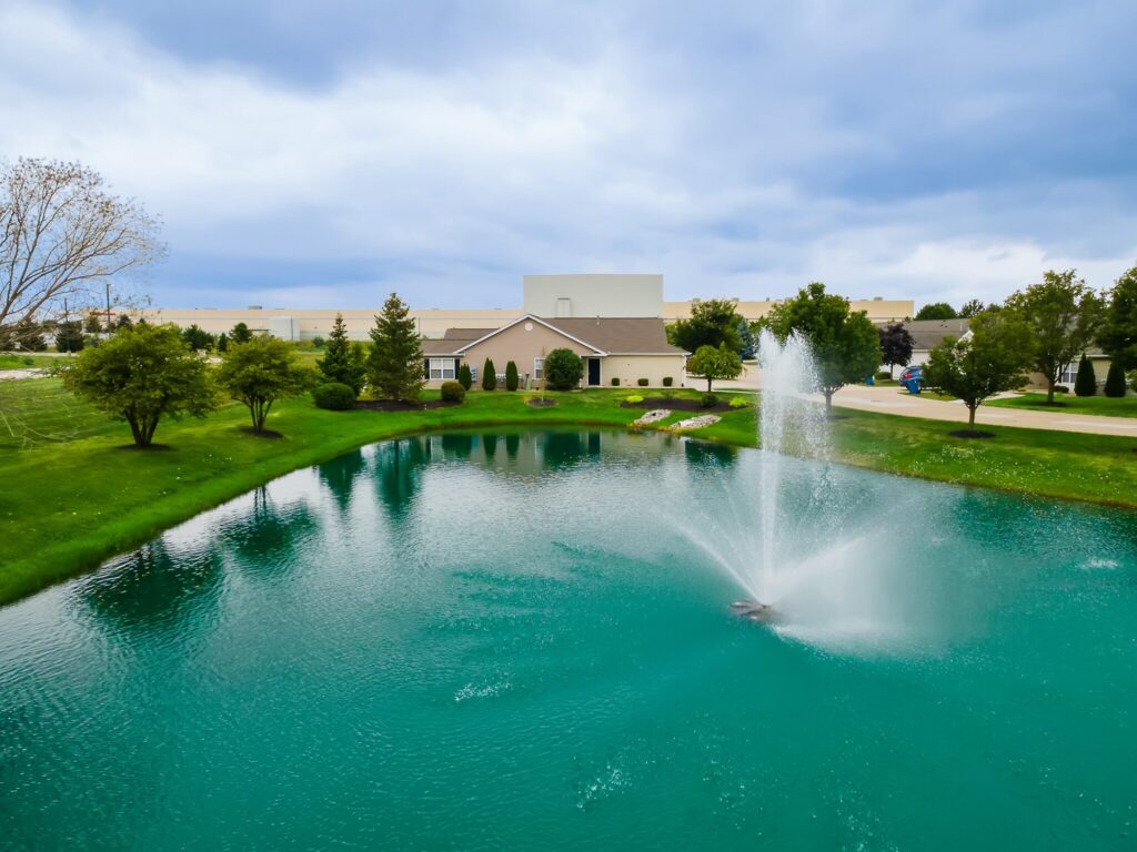A landscaped pond with a water fountain in front of a building, surrounded by green grass and trees under a cloudy sky.