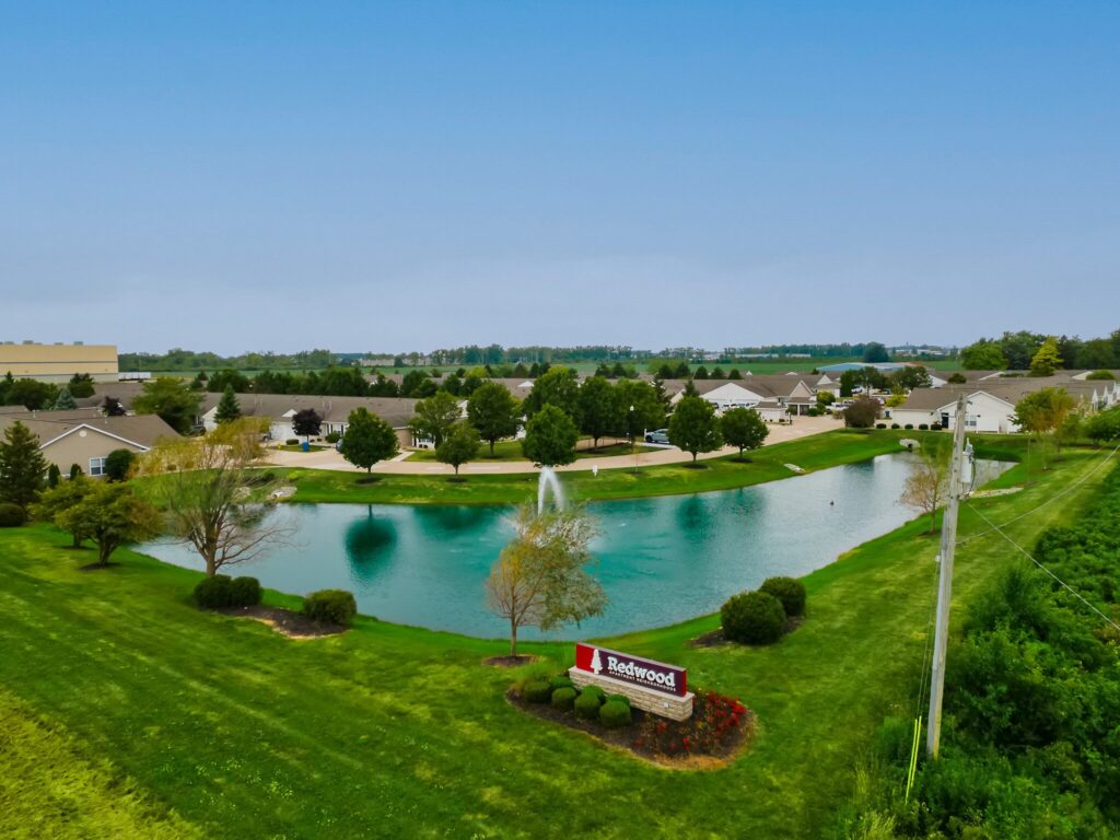 A landscaped pond with a fountain is surrounded by houses and trees. A sign reading "Redwood" is at the foreground. The sky is clear and blue.
