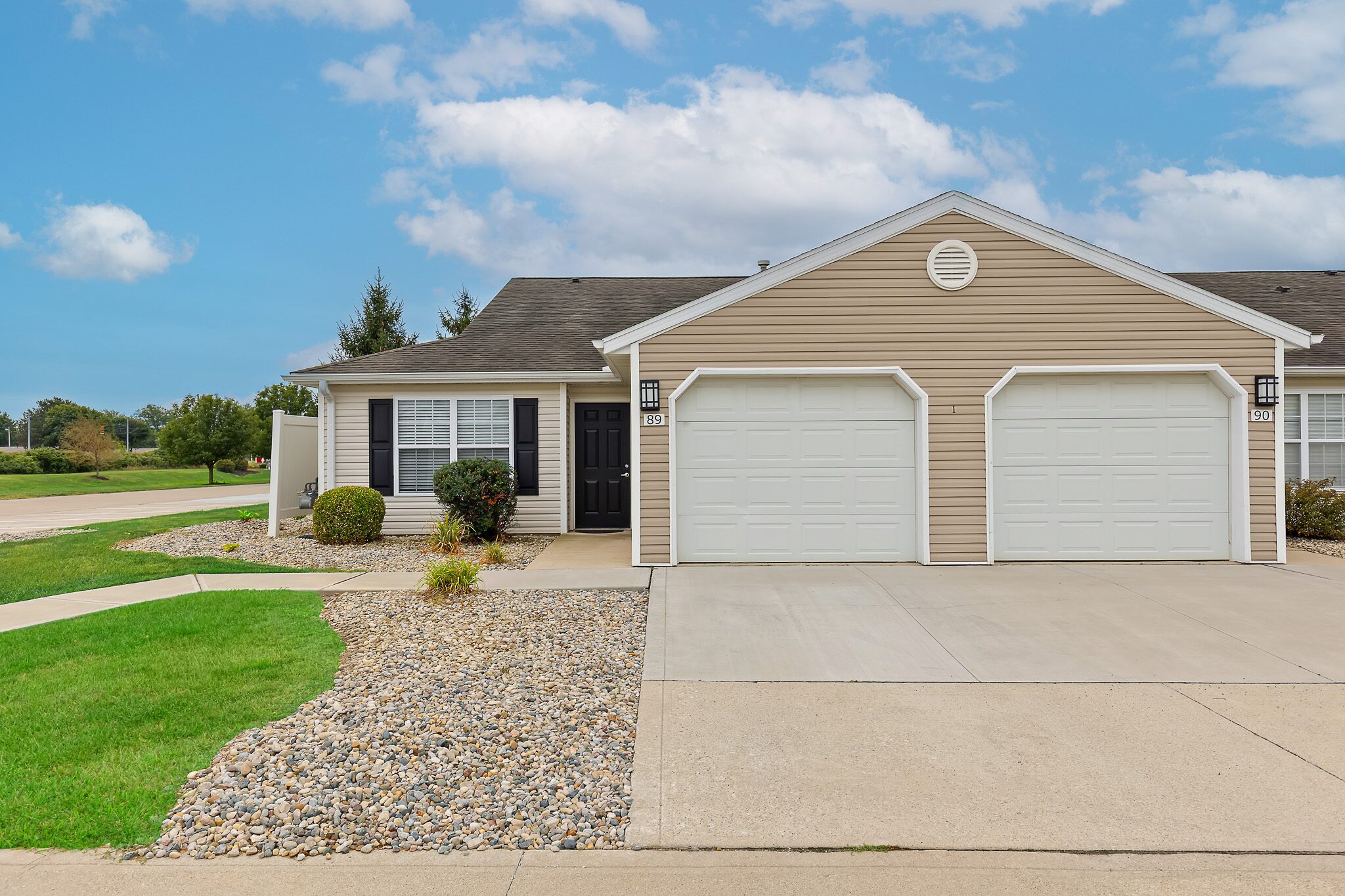 Single-story duplex with beige siding, two white garage doors, black front door, stone landscaping, and concrete driveway under a partly cloudy sky.