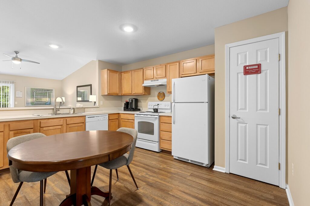 A kitchen with light wood cabinets, white appliances, a round dining table with four chairs, and a pantry door with a red sign. The floor is wood and walls are beige.
