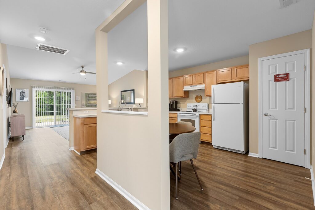 Open-concept kitchen and dining area with light wood cabinets, white appliances, and wood flooring, leading to a living room with sliding glass doors to a patio.