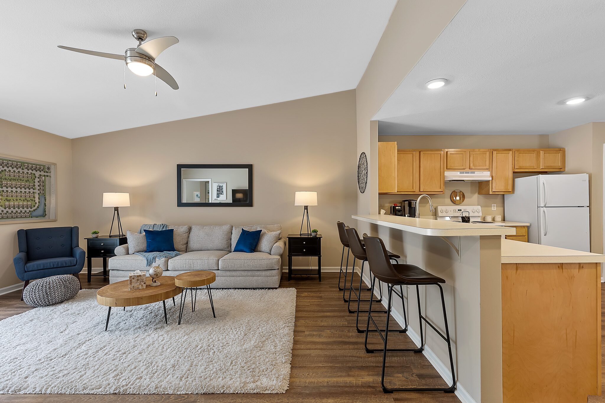 Modern open-concept living room and kitchen with neutral tones, a sofa, two tables, bar stools, and light wood cabinetry.