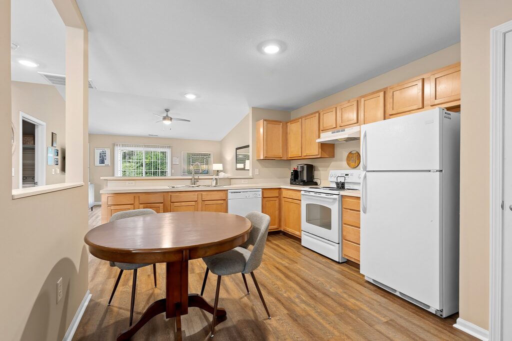 A kitchen with wooden cabinets, white appliances, a round wooden dining table with two chairs, and wood flooring. The space opens to a living area with large windows.