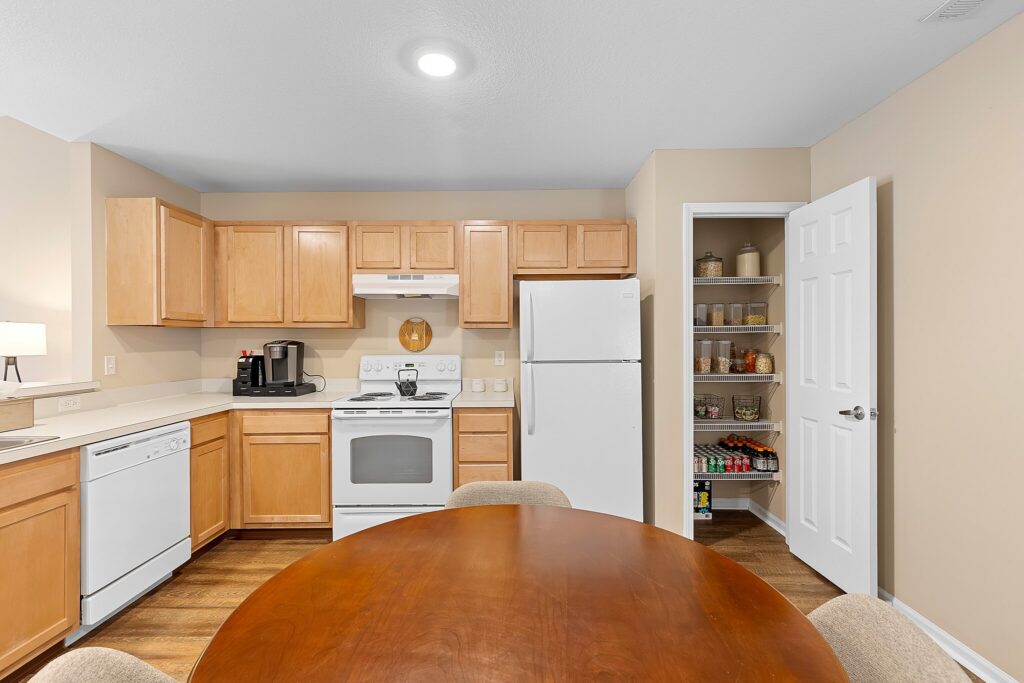 A kitchen with light wood cabinets, white appliances, a round wooden dining table, and an open pantry showing shelves with food items and containers.