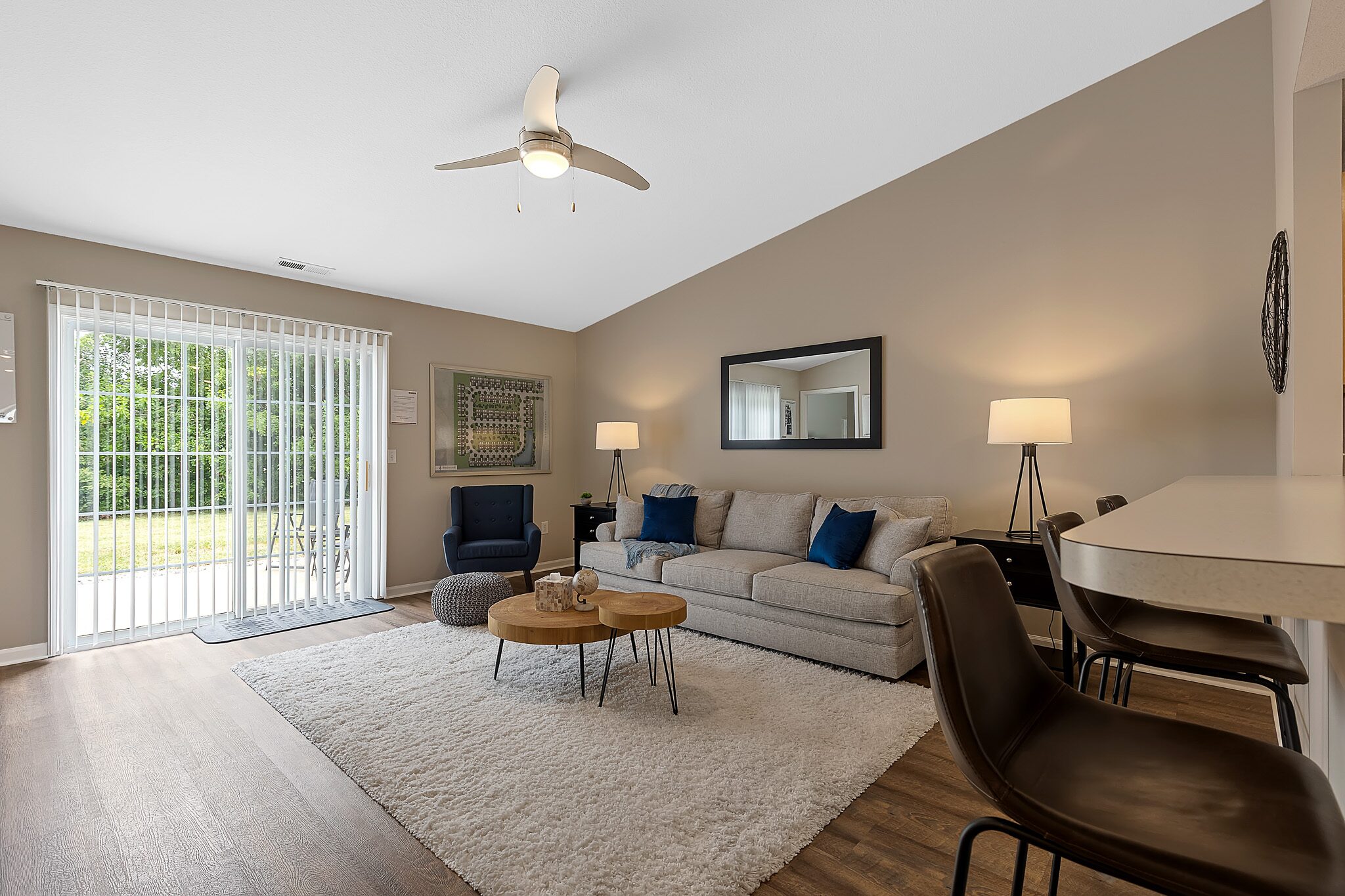 Modern living room with a beige sofa, armchair, white rug, coffee tables, bar stools, and large window with vertical blinds letting in natural light.