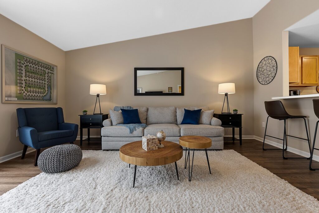 A modern living room with a gray sofa, two blue chairs, round wooden tables, a white rug, wall art, and two floor lamps, with a kitchen area visible on the right.