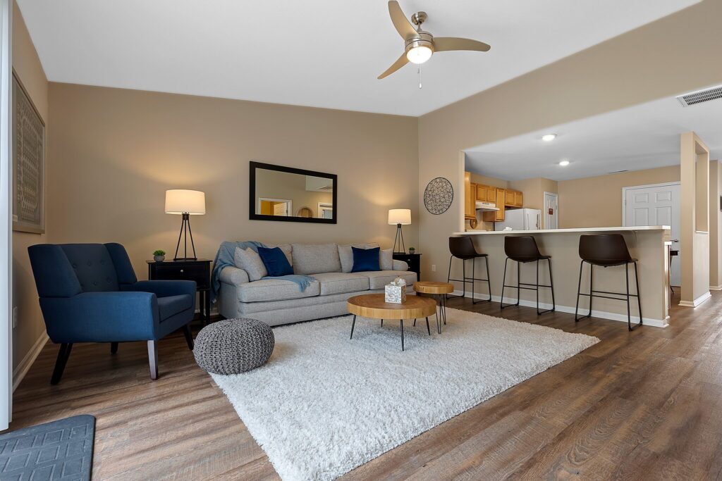 Modern living room with a sofa, armchair, coffee tables, and bar stools at a kitchen counter. Neutral tones, wood flooring, and contemporary decor create a cozy, open space.