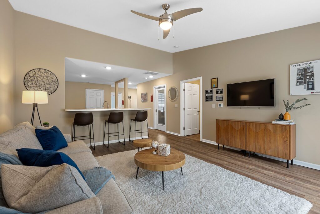 Modern living room with beige walls, a gray sofa, round wooden coffee table, wall-mounted TV, and open kitchen with bar stools in the background.
