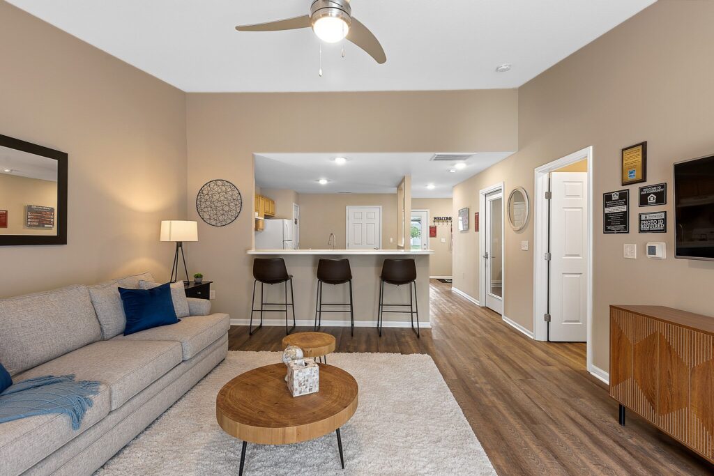 Modern living room with a gray sofa, round wooden coffee tables, a bar counter with three stools, and an open kitchen in the background. Wood flooring and neutral-colored walls.
