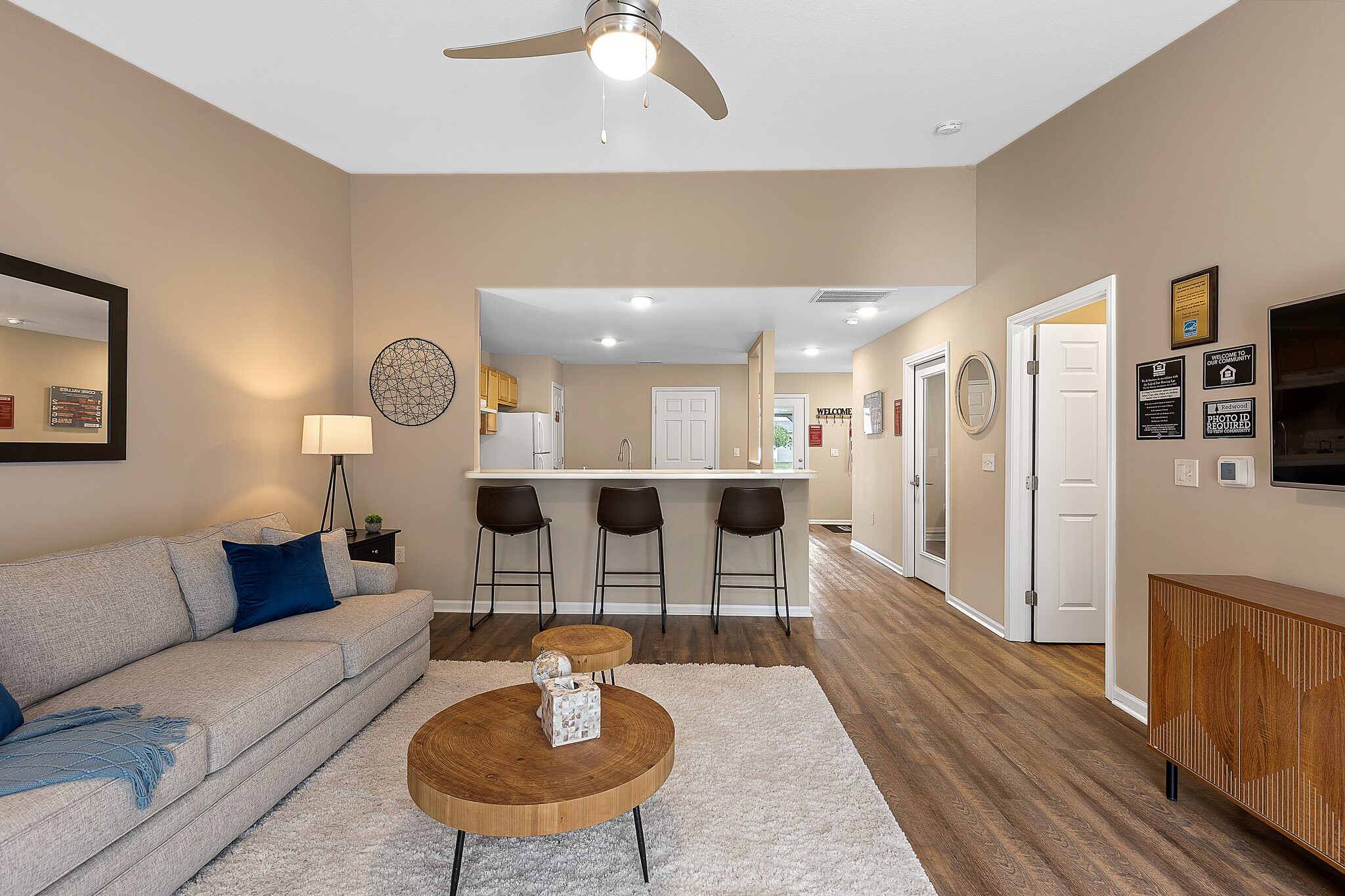 Modern living room with a gray sofa, round wooden coffee tables, a bar counter with three stools, and an open kitchen in the background. Wood flooring and neutral-colored walls.