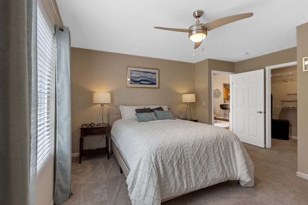A neatly made bed with white bedding in a beige bedroom, two bedside tables with lamps, a ceiling fan, and an open door leading to a walk-in closet.