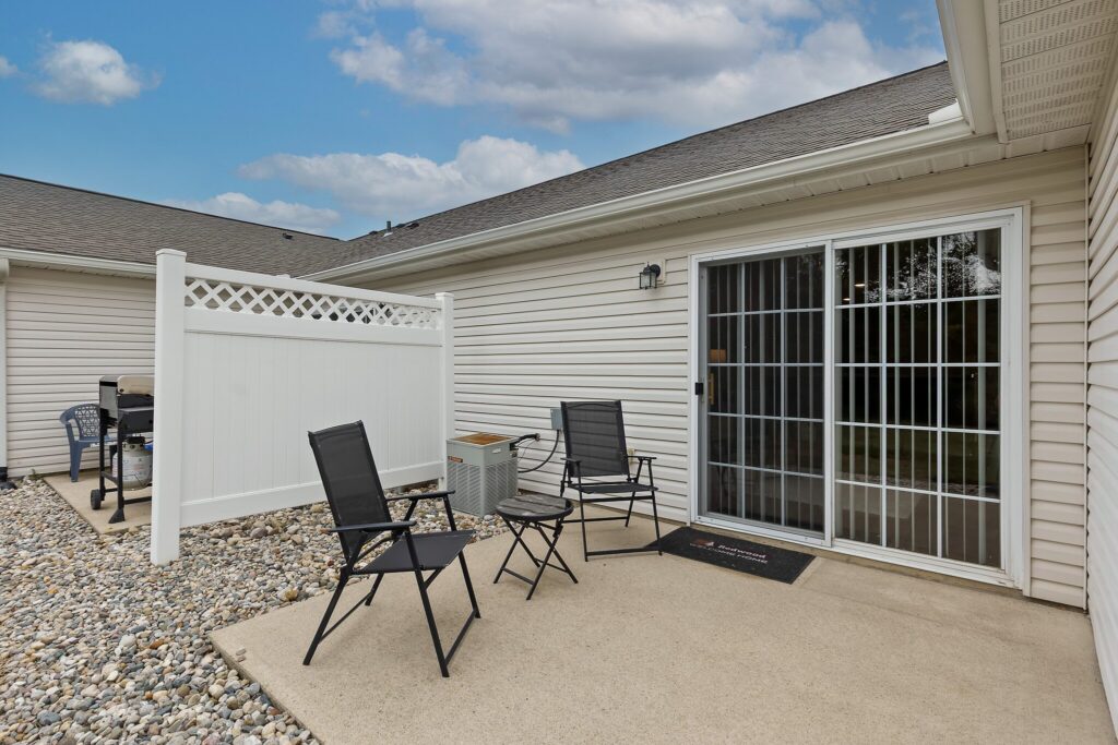 Outdoor patio with two black chairs and a small table on concrete, next to a sliding glass door. White privacy fence and rocks line the edge; grill is visible in the background.