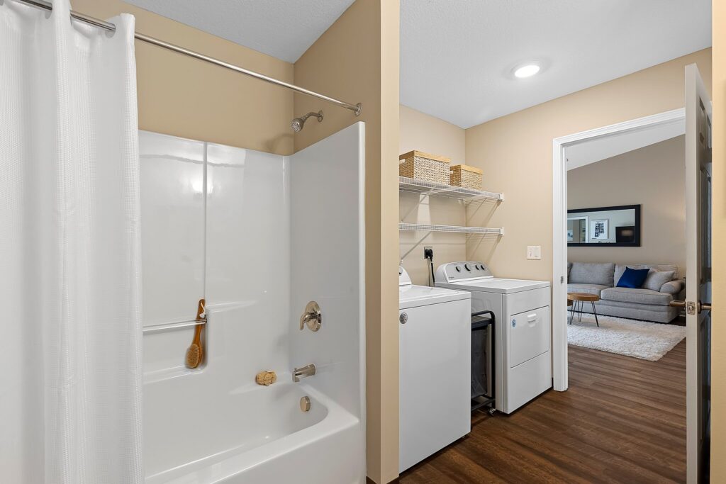 Bathroom and laundry area with a white bathtub, shower curtain, washing machine, dryer, shelves with baskets, and view into a living room with a sofa.