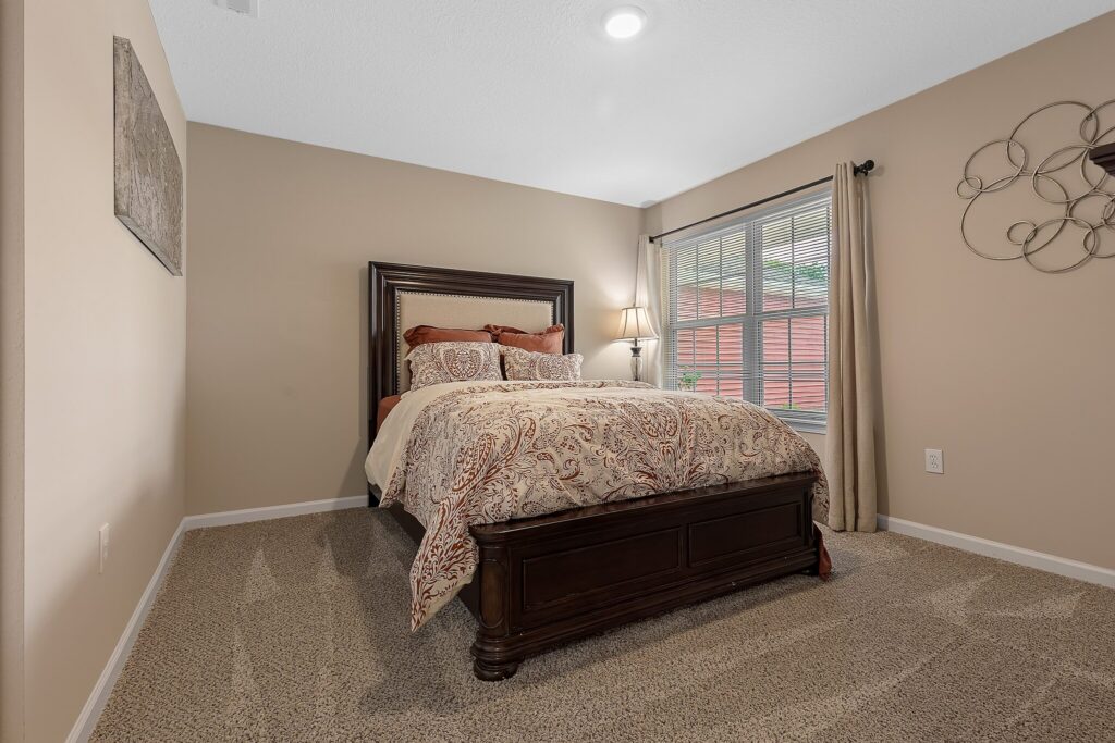 A neatly made bed with patterned bedding is centered in a carpeted bedroom with beige walls, a large window, and minimal decor.