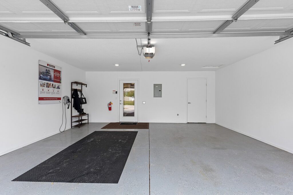 Clean, empty two-car garage with white walls, a door to the outside, coat hooks, a large wall poster, fire extinguisher, and rubber mats on the floor.